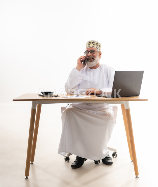 ايماءات السعادة والسرور, استخدام الأجهزة الحديثة والتقنيات المتطورة، مفهوم ادارة الاعمال عن بعد، مهن ووظائف مكتبيه عمانية, بورتريه لرجل كبير السن عماني يرتدي الدشداشة والكمه العمانية التقليدية يقوم باجراء مكالمة هاتفية عبر الهاتف المحمول, خلفية بيضاء