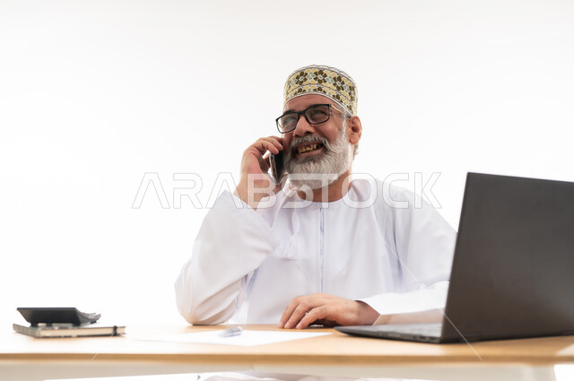 Gestures of happiness and pleasure, use of modern devices and advanced technologies, concept of remote business management, Omani office jobs and professions, close-up portrait of elderly Omani man wearing traditional Omani dishdasha and kummah making a phone call via mobile phone, white background