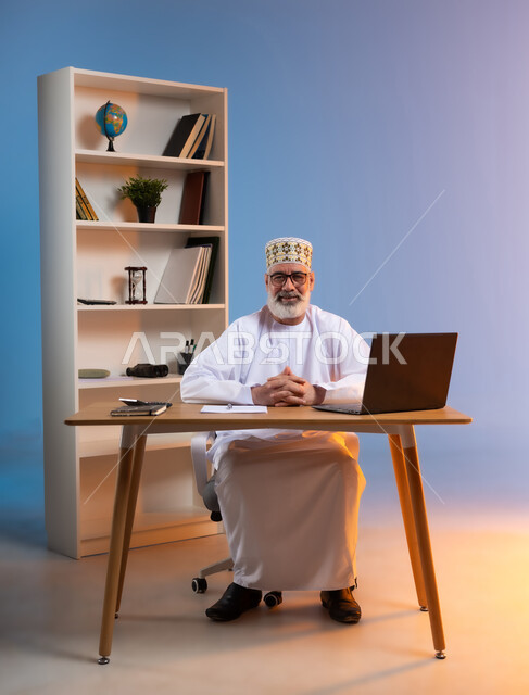 النظر الى الكاميرا بتعابير الثقة بالنفس, بورتريه لرجل كبير السن عربي خليجي عماني يرتدي الدشداشة والكمه العمانية التقليدية يجلس خلف طاولة خشبية بايماءات ايدي متشابكة, خلفية ملونة