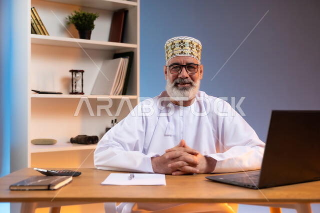 النظر الى الكاميرا بتعابير الثقة بالنفس, بورتريه مقرب لرجل كبير السن عربي خليجي عماني يرتدي الدشداشة والكمه العمانية التقليدية يجلس خلف طاولة خشبية بايماءات ايدي متشابكة, خلفية ملونة