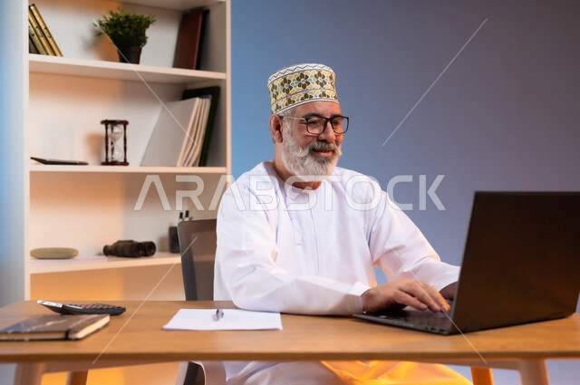 دمج التقنيات الحديثة والمتطورة في الحياة العملية، بورتريه لرجل كبير السن عربي خليجي عماني يرتدي الدشداشة والكمه العمانية التقليدية يجلس خلف طاولة خشبية ينظر الى جهاز الحاسوب المحمول بايماءات الفرح, خلفية ملونة