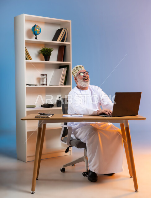 تعابير الضحك الشديد والسرور, استخدام التقنيات الحديثة والمتطورة في العمل، بورتريه لرجل كبير السن عربي خليجي عماني يرتدي الدشداشة والكمه العمانية التقليدية يجلس خلف طاولة خشبية يقوم بتبادل الدردشات عبر جهاز الحاسوب المحمول بايماءات الفرح, خلفية ملونة