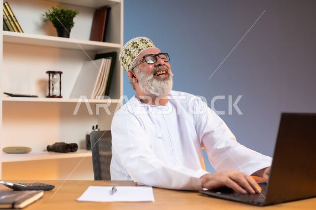 تعابير الضحك الشديد والسرور, استخدام التقنيات الحديثة والمتطورة في العمل، بورتريه مقرب لرجل كبير السن عربي خليجي عماني يرتدي الدشداشة والكمه العمانية التقليدية يجلس خلف طاولة خشبية يقوم بتبادل الدردشات عبر جهاز الحاسوب المحمول بايماءات الفرح, خلفية ملونة