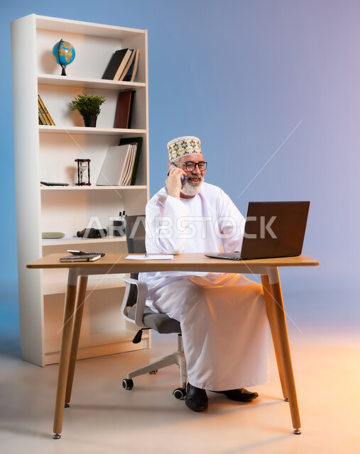 مفهوم ادارة الاعمال عن بعد، مهن ووظائف مكتبيه عمانية, استخدام الأجهزة الحديثة والتقنيات المتطورة، بورتريه لرجل عربي خليجي عماني يرتدي الدشداشة والكمه العمانية التقليدية يقوم باجراء مكالمة هاتفية عبر الهاتف المحمول, خلفية ملونة