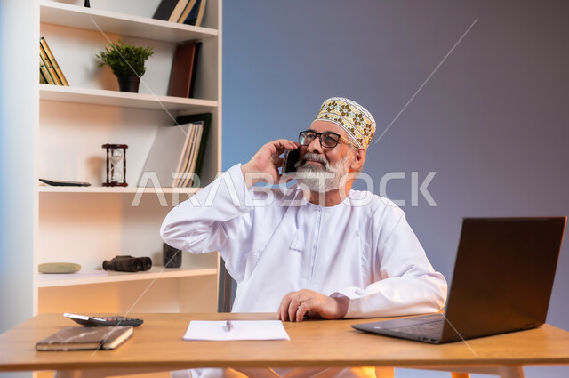 Using modern devices and advanced technologies, the concept of remote business management, Omani office jobs and professions, portrait of an Arab Gulf Omani man wearing a dishdasha and traditional Omani kummah making a phone call via mobile phone, colorful background