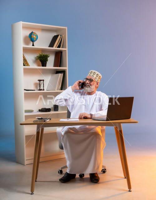 مفهوم ادارة الاعمال عن بعد، مهن ووظائف مكتبيه عمانية, استخدام الأجهزة الحديثة والتقنيات المتطورة، بورتريه لرجل عربي خليجي عماني يرتدي الدشداشة والكمه العمانية التقليدية يقوم باجراء مكالمة هاتفية عبر الهاتف المحمول, خلفية ملونة
