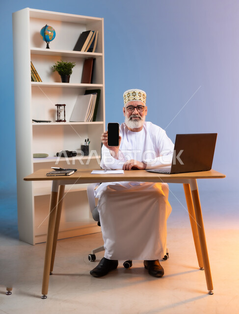 استخدام الأجهزة والتقنيات الحديثة، مهن و أعمال إدارية مكتبية، بورتريه لرجل كبير السن عربي خليجي عماني يرتدي الدشداشة والكمه يجلس خلف طاولة خشبية يقوم بعرض شاشة سوداء فارغة عبر الهاتف المحمول, النظر الى الكاميرا بتعابير السرور, خلفية ملونة