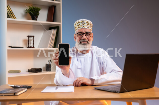 Using modern devices and technologies, office administrative professions and work, close-up portrait of an elderly Arab Gulf Omani man wearing a dishdasha and a kummah sitting behind a wooden table displaying a blank black screen via a mobile phone, looking at the camera with an expression of pleasure, colorful background