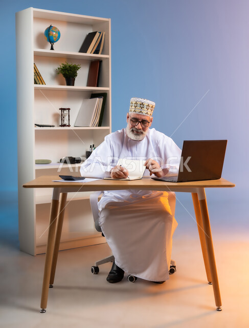 مهن ووظائف مكتبيه عمانية, مفهوم ادارة الاعمال عن بعد، بورتريه لرجل كبير السن عربي خليجي عماني يرتدي الدشداشة والكمه العمانية التقليدية يجلس خلف طاولة خشبية يقوم بتدوين الملاحظات, استخدام التقنيات الحديثة المتطورة, خلفية ملونة