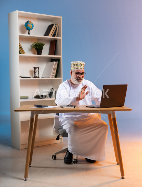 إجراء مكالمة فيديو عبر جهاز الحاسوب المحمول، استخدام التقنيات الحديثة في التواصل مع العملاء، بورتريه لرجل كبير السن عربي خليجي عماني يرتدي الدشداشة والكمه العمانية التقليدية يرفع يديه بايماءات القاء التحية والسلام, خلفية ملونة