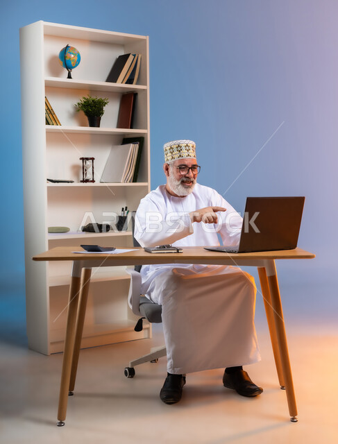 استخدام التقنيات الحديثة والمتطورة في العمل، بورتريه لرجل كبير السن عربي خليجي عماني يرتدي الدشداشة والكمه العمانية يجلس خلف طاولة خشبية يشير باصبع السبابة الى جهاز الحاسوب المحمول بايماءات السعادة والسرور, خلفية ملونة