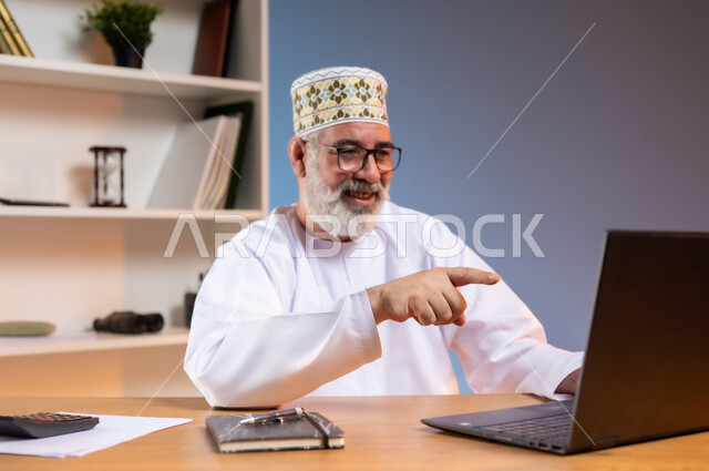 استخدام التقنيات الحديثة والمتطورة في العمل، بورتريه مقرب لرجل كبير السن عربي خليجي عماني يرتدي الدشداشة والكمه العمانية يجلس خلف طاولة خشبية يشير باصبع السبابة الى جهاز الحاسوب المحمول بايماءات السعادة والسرور, خلفية ملونة