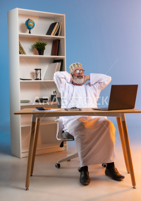 الراحه والاسترخاء بعد التعب, بورتريه لرجل كبير السن عربي خليجي عماني يرتدي الدشداشة والكمه العمانية التقليدية يجلس خلف طاولة خشبية يضع يديه خلف رقبته بايماءات التامل والتفكير في شيء ما, تعابير السرور والرضا عن العمل, خلفية ملونة