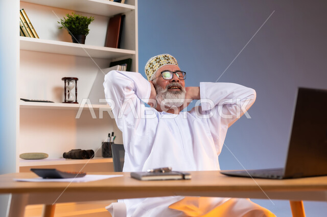 الراحه والاسترخاء بعد التعب, بورتريه مقرب لرجل كبير السن عربي خليجي عماني يرتدي الدشداشة والكمه العمانية التقليدية يجلس خلف طاولة خشبية يضع يديه خلف رقبته بايماءات التامل والتفكير في شيء ما, تعابير السرور والرضا عن العمل, خلفية ملونة