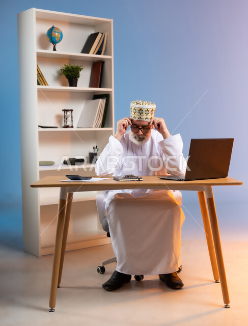 Expressions of fatigue and exhaustion, concept of remote business management, portrait of an elderly Arab Gulf Omani man wearing the traditional Omani dishdasha and kummah sitting behind a wooden table with his hands on his head in gestures of drowsiness and need for rest, colorful background