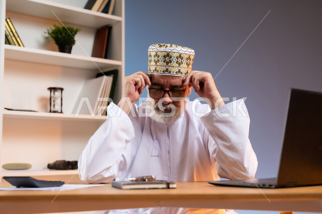 تعابير التعب والارهاق, مفهوم ادارة الاعمال عن بعد، بورتريه مقرب لرجل كبير السن عربي خليجي عماني يرتدي الدشداشة والكمه العمانية التقليدية يجلس خلف طاولة خشبية يضع يديه على رأسه بايماءات النعاس والحاجة الى الراحة, خلفية ملونة