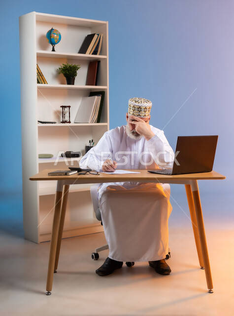 مهن ووظائف مكتبيه عمانية, مفهوم ادارة الاعمال عن بعد، بورتريه لرجل كبير السن عربي خليجي عماني يرتدي الدشداشة والكمه العمانية التقليدية يجلس خلف طاولة خشبية يضع يده على رأسه بتعابير التعب والارهاق, تدوين الملاحظات والافكار المهمة, خلفية ملونة