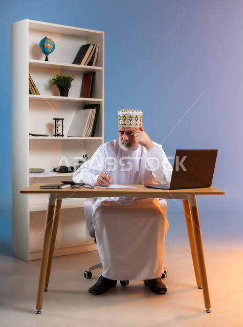 مهن ووظائف مكتبيه عمانية, مفهوم ادارة الاعمال عن بعد، بورتريه لرجل عربي خليجي عماني يرتدي الدشداشة والكمه العمانية التقليدية يجلس خلف طاولة خشبية يقوم بتدوين الملاحظات بايماءات التعب والارهاق, تعابير النعاس والحاجة الى الراحة, خلفية ملونة