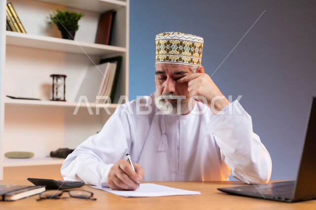 مهن ووظائف مكتبيه عمانية, مفهوم ادارة الاعمال عن بعد، بورتريه مقرب لرجل عربي خليجي عماني يرتدي الدشداشة والكمه العمانية التقليدية يجلس خلف طاولة خشبية يقوم بتدوين الملاحظات بايماءات التعب والارهاق, تعابير النعاس والحاجة الى الراحة, خلفية ملونة