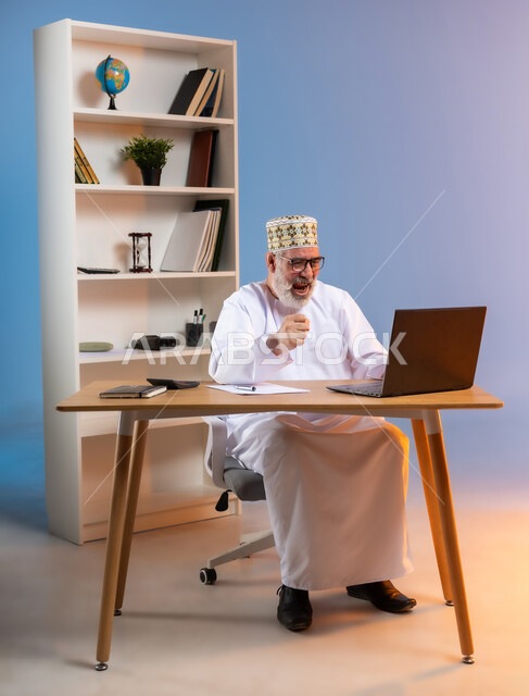 الفرح بالإنجاز وتحقيق الأهداف، بورتريه لرجل كبير بالسن عربي خليجي عماني يرتدي الدشداشة والكمه العمانية التقليدية والنظارة الطبية يجلس خلف طاولة خشبية ينظر الى الحاسوب المحمول بايماءات النصر والفوز, خلفية ملونة