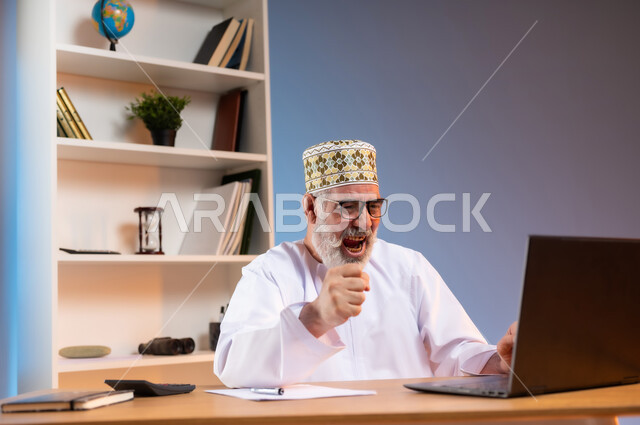 الفرح بالإنجاز وتحقيق الأهداف، بورتريه مقرب لرجل كبير بالسن عربي خليجي عماني يرتدي الدشداشة والكمه العمانية التقليدية والنظارة الطبية يجلس خلف طاولة خشبية ينظر الى الحاسوب المحمول بايماءات النصر والفوز, خلفية ملونة