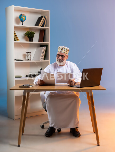مفهوم ادارة الاعمال عن بعد، مهن ووظائف مكتبيه، بورتريه لرجل كبير السن عربي خليجي عماني يرتدي الدشداشة والكمه يجلس خلف طاولة خشبية يحمل بيده الاوراق ويقوم بتدقيق العمل بتعابير الرضا والسرور, خلفية ملونة