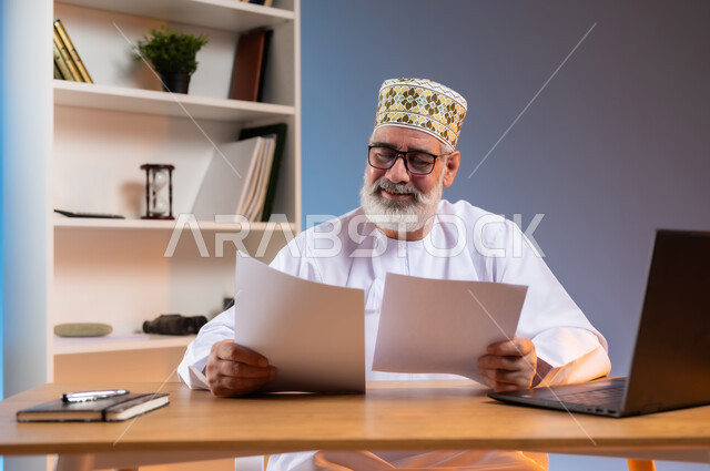 مفهوم ادارة الاعمال عن بعد، مهن ووظائف مكتبيه، بورتريه مقرب لرجل كبير السن عربي خليجي عماني يرتدي الدشداشة والكمه يجلس خلف طاولة خشبية يحمل بيده الاوراق ويقوم بتدقيق العمل بتعابير الرضا والسرور, خلفية ملونة