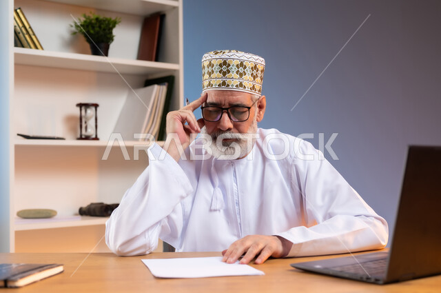 استخدام التقنيات الحديثة المتطورة, بورتريه مقرب لرجل كبير السن عربي خليجي عماني يرتدي الكمه والدشداشة يضع يده على رأسه بتعابير التفكير و التذكر, محاولة ايجاد حل لمشكلة ما, تعابير شرود الذهن و التركيز و التمعن, خلفية ملونة