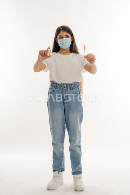 مفهوم الوعي الصحي، تجنب إنتشار العدوى، التطعيم الآمن لكوفيد19، اتباع اجراءات وزارة الصحة الوقائية، بورتريه لفتاة عربية خليجية إماراتية ترتدي الكمامة تحمل بيدها ابرة وجرعة من لقاح فايروس كورونا، الحماية من الأمراض والحفاظ على السلامة، الجسم الكامل بالطول، خلفية بيضاء