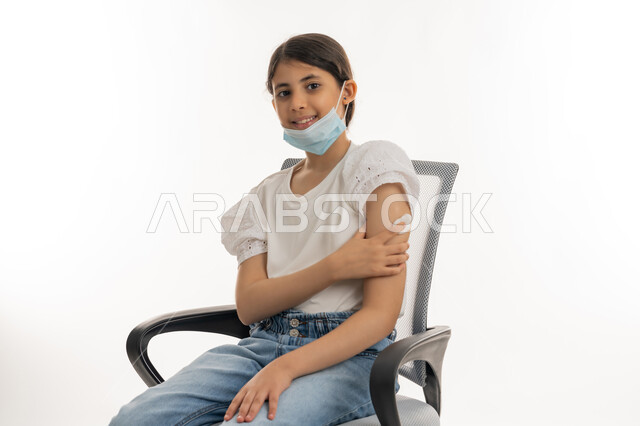 النظر الى الكاميرا بإيماءات القوة والسرور، تجنب إنتشار العدوى، الحماية من الأمراض والحفاظ على السلامة، التطعيم الآمن لكوفيد19، اتباع اجراءات وزارة الصحة الوقائية، مفهوم الوعي الصحي، بورتريه لفتاة عربية خليجية إماراتية مبتسمة ترتدي الكمامة تضع يدها مكان الحقن، خلفية بيضاء