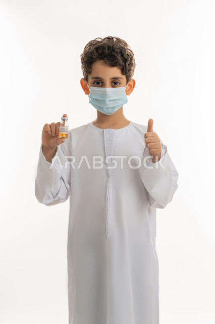 رفع اصبع الإبهام للاعجاب والقبول، اتباع اجراءات وزارة الصحة الوقائية، مفهوم الوعي الصحي، الحماية من الأمراض والحفاظ على السلامة، التطعيم الآمن لكوفيد19، بورتريه لولد عربي خليجي إماراتي يرتدي الكندورة والكمامة يحمل بيده جرعة من لقاح فايروس كورونا، تجنب انتشار العدوى، خلفية بيضاء