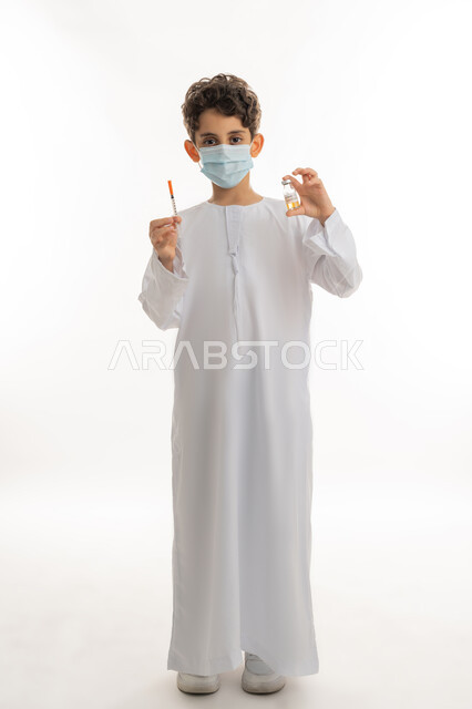 Following the preventive measures of the Ministry of Health, the concept of health awareness, protection from diseases and maintaining safety, safe vaccination for Covid-19, avoiding the spread of infection, portrait of an Arab Gulf Emirati boy wearing a kandura and a mask holding a needle and a dose of the Corona virus vaccine in his hand, full body length, white background
