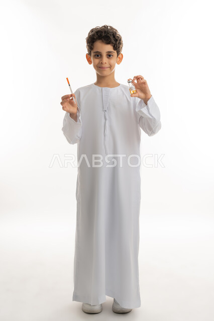 Protection from diseases and maintaining safety, avoiding the spread of infection, the concept of health awareness, a portrait of an Arab Gulf Emirati boy wearing a kandura and a mask, holding a needle and a dose of the Corona virus vaccine in his hand, following preventive measures, safe vaccination for Covid-19, full body length, white background