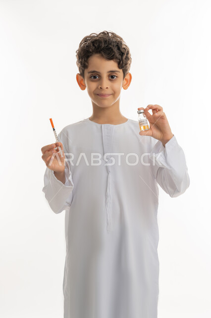 التطعيم الآمن لكوفيد19، مفهوم الوعي الصحي، الحماية من الأمراض والحفاظ على السلامة، تجنب إنتشار العدوى، بورتريه لولد عربي خليجي إماراتي يرتدي الكندورة يحمل بيده ابرة وجرعة من لقاح فايروس كورونا، اتباع اجراءات الوقاية ، النظر الى الكاميرا بتعابير الفرح والسرور، خلفية بيضاء