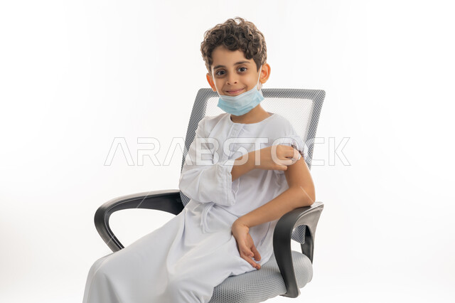 Health awareness concept, avoiding the spread of infection, protecting against diseases and maintaining safety, portrait of an Arab Gulf Emirati boy wearing a kandura and a mask sitting on a chair, following preventive measures, safe vaccination for Covid-19, white background
