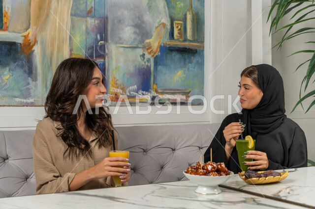 بناء علاقات اجتماعية ناجحة، النقاشات والحوارات في الأجواء الهادئة، قضاء أوقات ممتعة ومسلية مع الأصدقاء، صورة مقربة لإمرأة عربية خليجية اماراتية ترتدي الحجاب والعباءة السوداء تجلس على الأريكة مع صديقتها بإحدى المقاهي بإيماءات السعادة، مشاركة المشروبات الباردة