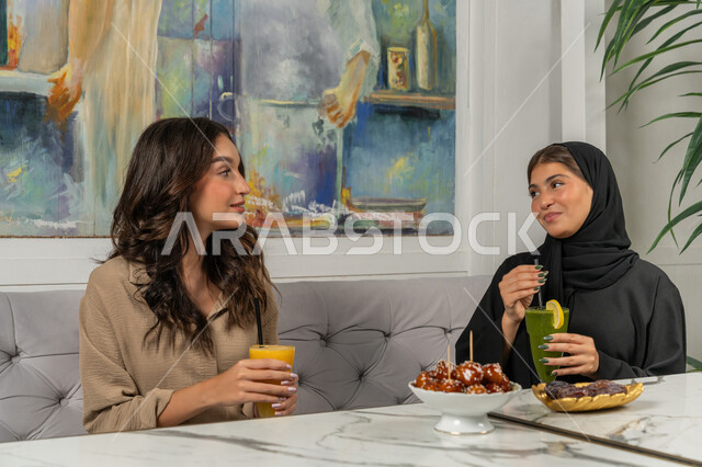 النقاشات والحوارات في الأجواء الهادئة، بناء علاقات اجتماعية ناجحة، قضاء أوقات ممتعة ومسلية مع الأصدقاء، صورة مقربة لإمرأة عربية خليجية اماراتية ترتدي الحجاب والعباءة السوداء تجلس على الأريكة مع صديقتها بإحدى المقاهي بإيماءات السعادة، مشاركة المشروبات الباردة