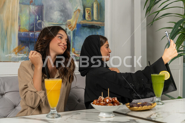 توثيق اللحظات السعيدة من خلال التقاط الصور التذكارية، استخدام اتكنولوجيا والتقنيات الحديثة، صورة مقربة لإمرأة عربية خليجية اماراتية ترتدي الحجاب والعباءة السوداء تجلس على الأريكة مع صديقتها بإحدى المقاهي وتستخدم الهاتف، مشاركة المشروبات الباردة والحلويات