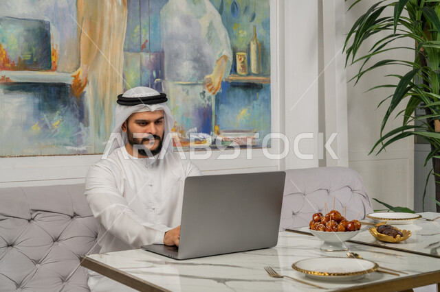 إدارة شؤون الأعمال وتنظيمها عبر الأجهزة التقنية الحديثة، استخدام التكنولوجيا والتقنيات المتطورة، إنجاز مهام العمل عن بعد، صورة مقربة لرجل عربي خليجي اماراتي يرتدي الكندورة والغترة البيضاء يجلس على الأريكة في إحدى المقاهي الراقية ويستخدم الحاسوب المحمول