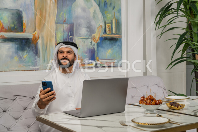 Using advanced technology and techniques, accomplishing work tasks remotely, managing and organizing business affairs through modern technical devices, a close-up image of an Emirati Gulf Arab man wearing a white kandura and ghutra sitting on the couch in an upscale cafe and using a phone and laptop