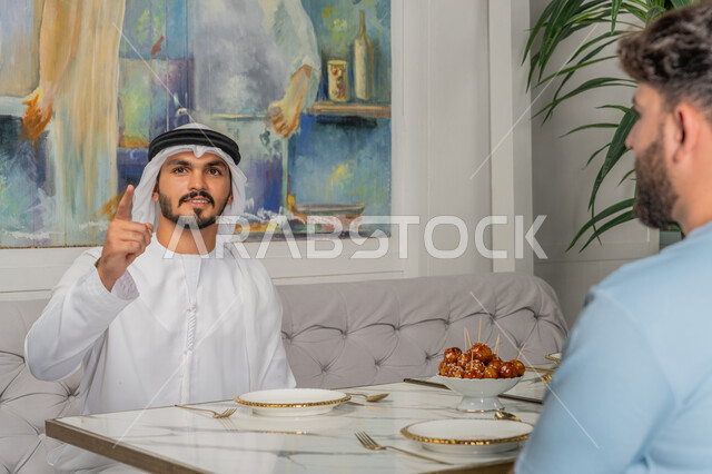 تعابير السعادة والسرور، الاستمتاع في وقت الفراغ مع الأصدقاء، علاقات اجتماعية ناجحة بين الأشخاص، صورة مقربة لرجل عربي خليجي اماراتي يرتدي الكندورة والغترة البيضاء يجلس على الأريكة مع صديقه في إحدى المقاهي الراقية ويشير بإصبع السبابة الى شيء ما والنظر لمكان معين