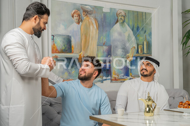 بناء علاقات اجتماعية ناجحة بين الأشخاص، قضاء أوقات ممتعة مع الأصدقاء، صورة مقربة لرجل عربي خليجي اماراتي يرتدي الكندورة والغترة البيضاء يجلس على الأريكة مع أصدقائه في إحدى المقاهي الراقية بتعابير السعادة والسرور، شاب يقف ويمسك بيد صديقه بإيماءات القوة والشجاعة