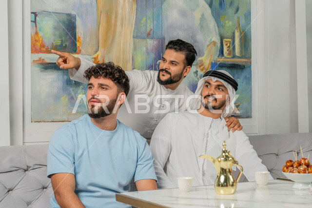 قضاء أوقات ممتعة مع الأصدقاء، علاقات اجتماعية ناجحة بين الأشخاص، صورة مقربة لرجل عربي خليجي اماراتي يرتدي الكندورة والغترة البيضاء يجلس على الأريكة مع أصدقائه في إحدى المقاهي الراقية بتعابير السعادة والسرور، شاب يشير بإصبع السبابة الى شيء ما والنظر لمكان معين