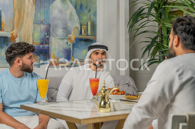 النقاشات والحوارات في الأجواء الهادئة، مشاركة المشروبات الباردة والقهوة، علاقات اجتماعية ناجحة بين الأشخاص، قضاء أوقات ممتعة مع الأصدقاء، رجل عربي خليجي اماراتي يرتدي الكندورة والغترة البيضاء يجلس مع أصدقائه في إحدى المقاهي الراقية بتعابير السعادة والسرور
