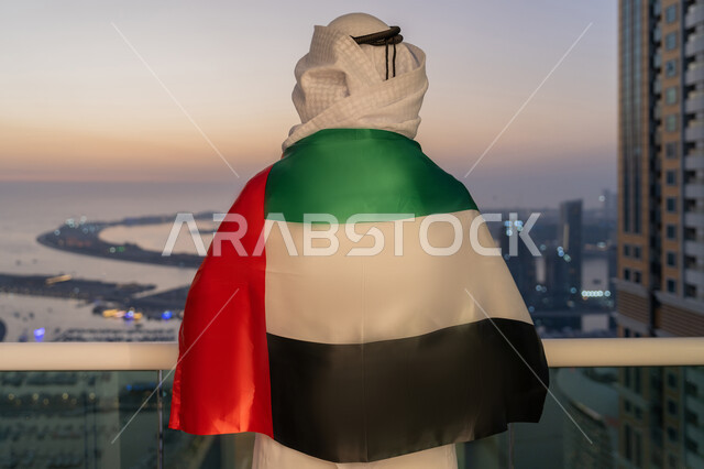 مفهوم حب الوطن والعلم، الإحتفال بالعيد الوطني الإماراتي 2 ديسمبر، إحياء ذكرى استقلال دولة الإمارات العربية المتحدة، صورة مقربة من الخلف لرجل عربي خليجي إماراتي يرتدي الكندورة والغترة يحمل علم الإمارات على ظهره ينظر الى الابراج، الاعتزاز والفخر بإنجازات الوطن