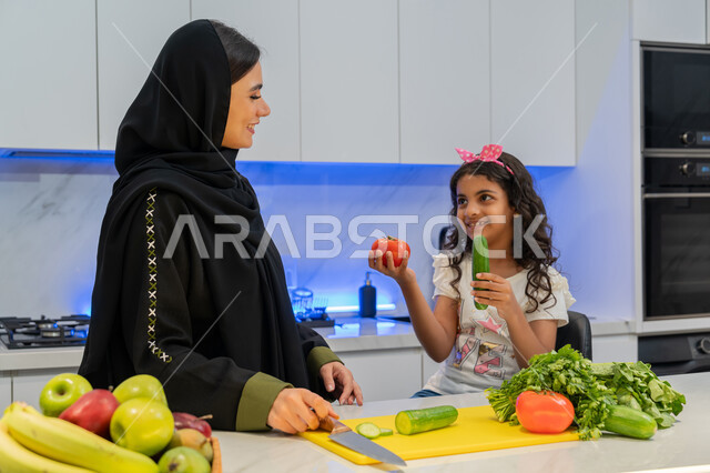 تعلم وصفات جديدة عبر الحاسوب المحمول، إعداد السلطات والأطباق المفيدة، المشاركة بالأعمال المنزلية، عائلة سعودية تقضي اوقات ممتعة، ام عربية خليجية اماراتية محجبة ترتدي العباءة تقف أمام طاولة عليها مجموعة من الخضراوات  في المطبخ مع ابنتها، اتباع نظام غذائي صحي