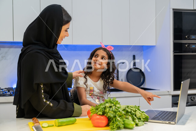إعداد السلطات والأطباق المفيدة، تعلم وصفات جديدة عبر الحاسوب المحمول،  المشاركة بالأعمال المنزلية، عائلة سعودية تقضي اوقات ممتعة، ام عربية خليجية اماراتية محجبة ترتدي العباءة تقف أمام طاولة عليها مجموعة من الخضراوات  في المطبخ مع ابنتها، اتباع نظام غذائي صحي