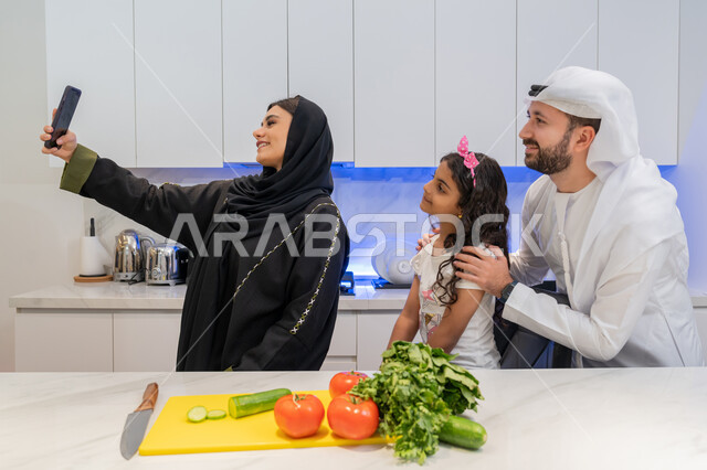 توثيق اللحظات التذكارية، التقاط صورة سيلفي عبر الهاتف المحمول، قضاء اوقات عائلية ممتعة، تحضير الوصفات والمأكولات الشهية، عائلة عربية خليجية اماراتية تقف أمام طاولة عليها مجموعة من الخضراوات  في المطبخ، اتباع نظام غذائي صحي، إعداد السلطات والأطباق المفيدة