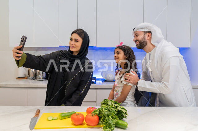 التقاط صورة سيلفي عبر الهاتف المحمول، توثيق اللحظات التذكارية، قضاء اوقات عائلية ممتعة، تحضير الوصفات والمأكولات الشهية، عائلة عربية خليجية اماراتية تقف أمام طاولة عليها مجموعة من الخضراوات  في المطبخ، اتباع نظام غذائي صحي، إعداد السلطات والأطباق المفيدة