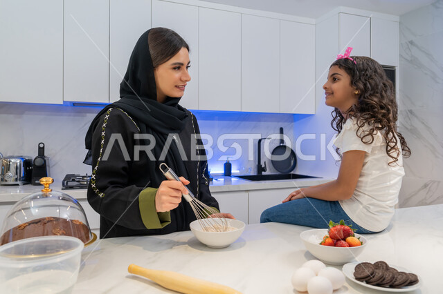 تعلم وصفات جديدة، قضاء أوقات ممتعه مع الابنه في المطبخ، اعداد وتحضير الحلويات الطازجه في المنزل, الوقوف أمام طاولة المطبخ, مقادير صنع الكعك، ام عربيه خليجية امارتية محجبه تقف مع ابنتها يقومان بتحضير الطعام، عائلة سعودية سعيدة، المشاركة بالأعمال المنزلية